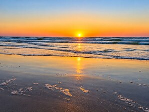 Ein Strand mit Fußabdrücken, die zum Wasser führen, Wellen und einem Sonnenuntergang.