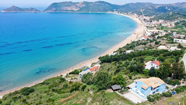 Vista aérea de uma vila costeira com uma praia, casas e montanhas ao fundo.