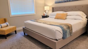 Een slaapkamer met een groot bed, een nachtkastje, een lamp, een stoel en een raam met jaloezieën.