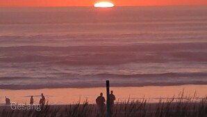 Een zonsondergang boven de oceaan met silhouetten van mensen en een paal op de voorgrond.