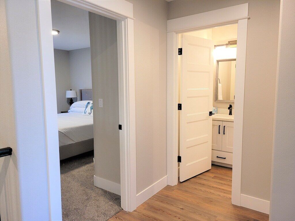 A hallway leading to a bedroom and a bathroom. The bedroom has a bed with white bedding and a nightstand with a lamp. The bathroom has a sink and a mirror.