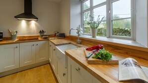 Une cuisine moderne avec des armoires blanches, un plan de travail en bois, une hotte noire, une fenêtre donnant sur un espace verdoyant et une planche à découper avec des légumes.