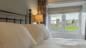 Un lit bien fait, avec du linge de lit blanc, une lampe décorée d’un motif de cerf et une fenêtre donnant sur une pelouse verdoyante et des bâtiments.
