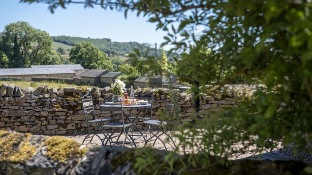 Un mur de pierre avec une table et des chaises disposées pour les repas, donnant sur un paysage rural avec des bâtiments et des arbres.