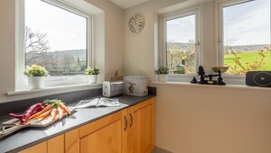 A kitchen with wooden cabinets, a black countertop, and a view of the countryside through large windows. - 1 Riverside Cottage, Burnsall, Yorkshire Dales (Burnsall)