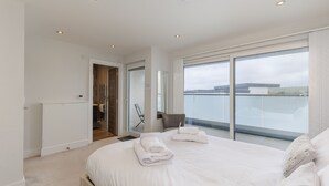 Ein Schlafzimmer mit einem großen Bett, einem Sessel, einem Spiegel und Blick auf ein Gewässer durch eine Schiebetür.