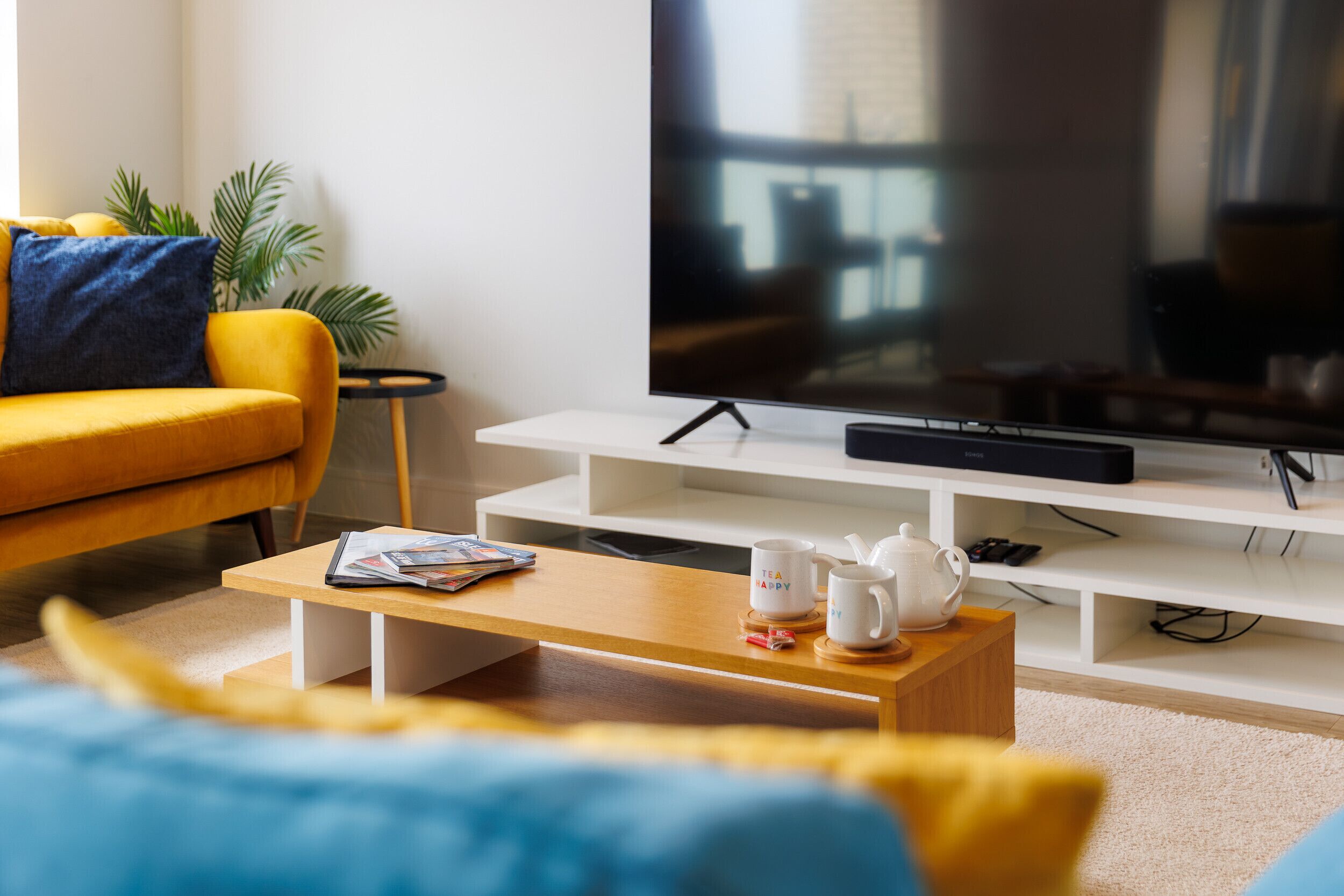 Un salon moderne avec un canapé jaune, une table basse sur laquelle sont posés des magazines et des tasses, un téléviseur à écran plat sur un meuble blanc et une plante dans un coin.