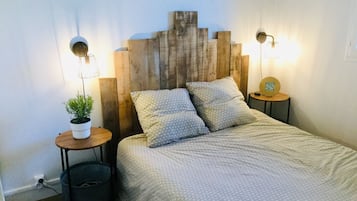 Une chambre avec une tête de lit en bois, un lit recouvert d’une literie à motifs, deux tables de chevet avec des lampes et une petite plante en pot.
