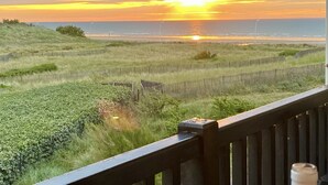 A sunset view from a balcony with a wooden railing, grassy fields, and a distant shoreline.