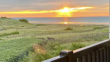 Une vue sur le coucher de soleil depuis un balcon doté d’une balustrade en bois, avec des champs verdoyants et une côte lointaine.