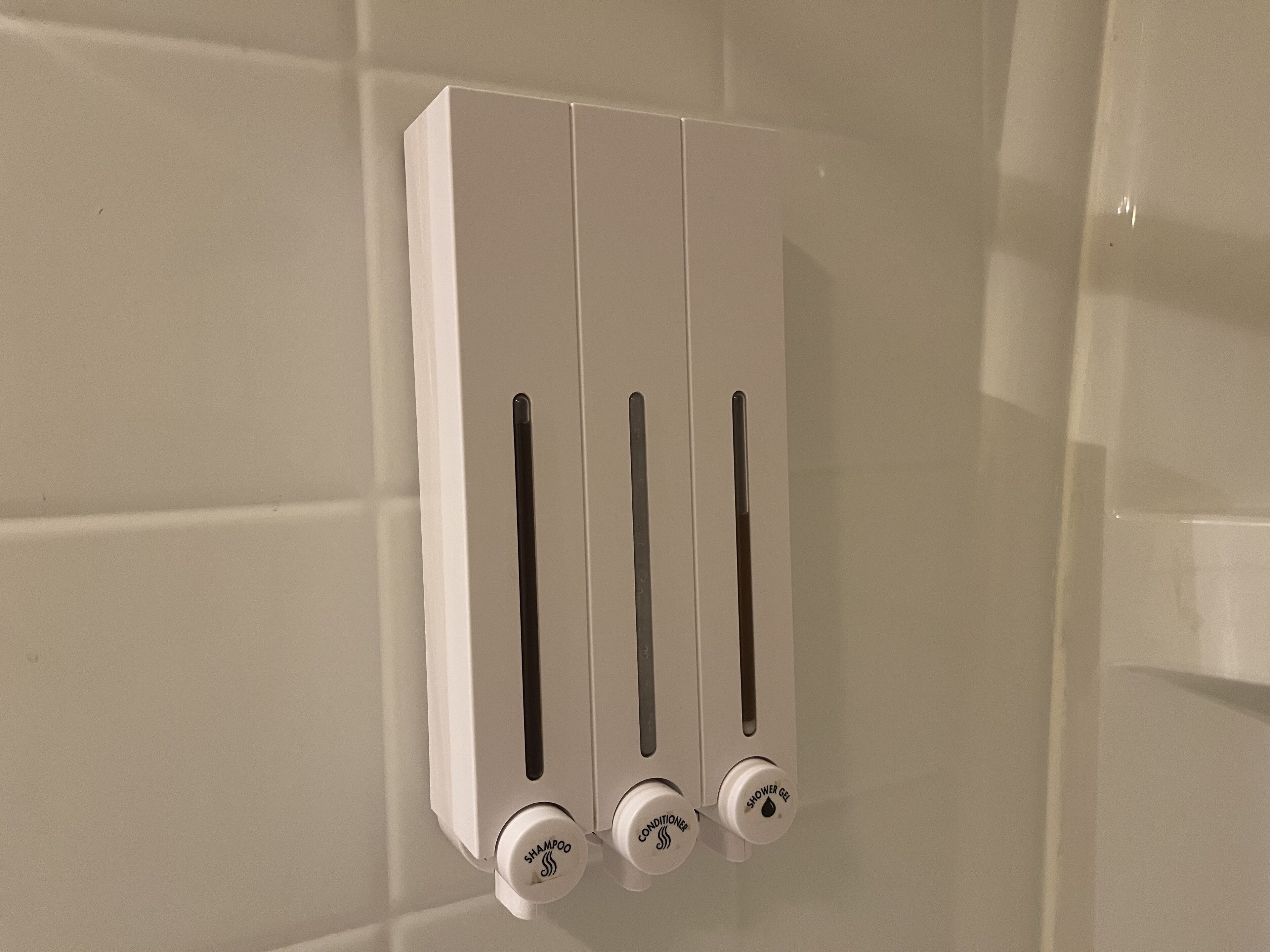 A wall-mounted dispenser with three slots for shampoo, conditioner, and shower gel.