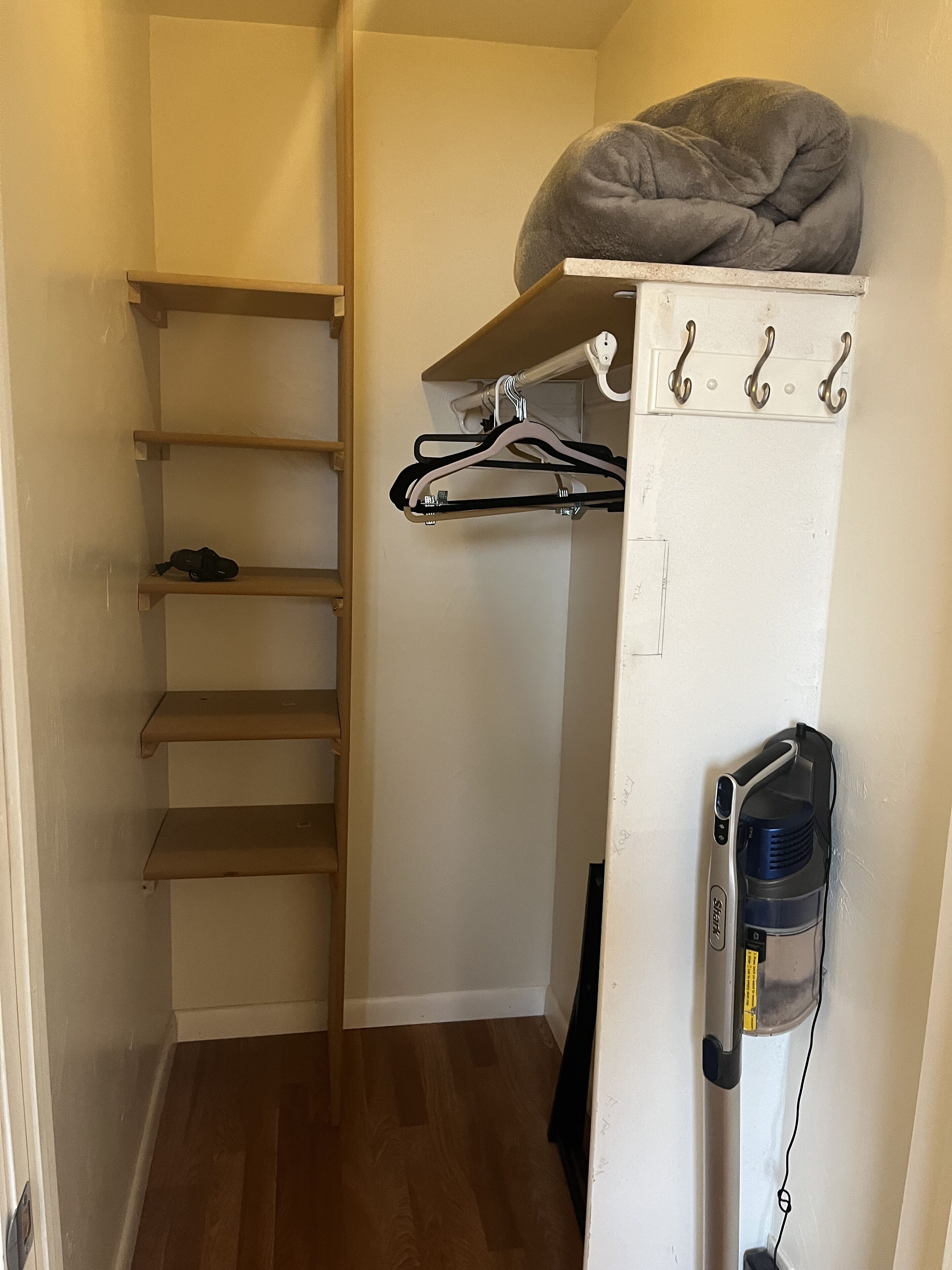 A compact room with a built-in closet, a grey pet bed on top, and a vacuum cleaner against the wall.