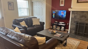 Ein Wohnzimmer mit einer Ledersofa, einem Couchtisch mit Dekoration, einem flachbildfernseher an der Wand, einem Kamin und einem Bücherregal mit Büchern und Dekogegenständen.