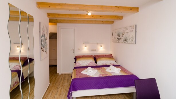 A bedroom with a bed, wooden beams, a mirror, and a wall-mounted light fixture. - Lavender Garden Apartments - Comfort Studio Apartment (Ground Floor) (Dubrovnik)