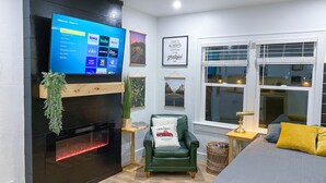 A modern living room with a flat-screen TV mounted on a black wall, a green armchair, a bed with a yellow pillow, and a fireplace with a red light. - Modern Studio in the Heart of Grants Pass (Grants Pass)