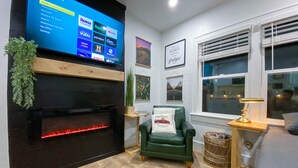 A modern living room with a fireplace, a television mounted on the wall, a green armchair, a small table with a lamp, and a window with blinds. - Modern Studio in the Heart of Grants Pass (Grants Pass)