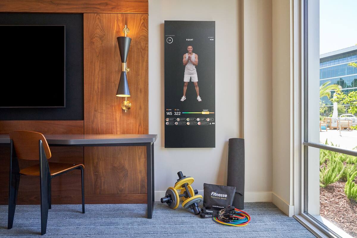 Ett hotellrum med ett skrivbord i trä, en stol, en tv, en affisch med en man som gör en knäböj och träningsutrustning.