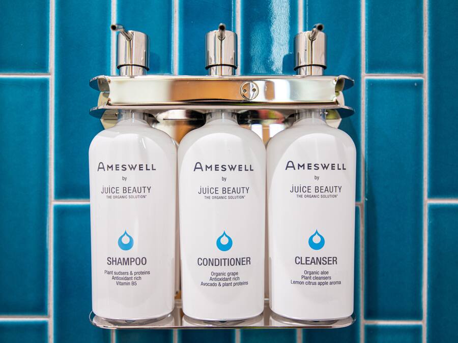 Tres productos de Amswell Juice Beauty: champú, acondicionador y limpiador, exhibidos en una estantería frente a una pared de azulejos azules.