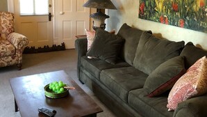 Una sala de estar con un sofá verde, una mesa de centro con un cuenco de manzanas verdes, una lámpara y un cuadro de flores en la pared.
