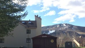 A building with a shuttle bus stop sign, a small wooden structure, and a mountain in the background. - Redecorated! Great Location at Mount Snow, Free Shuttle, Pool/Hot Tub (West Dover)