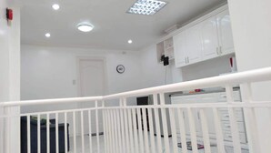Ruime kamer met een wit plafond, inbouwverlichting, een witte balustrade en uitzicht op een keuken met witte kasten.