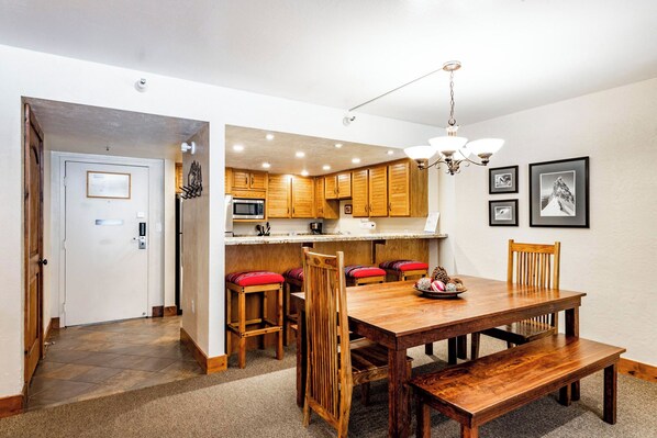 A dining area with a wooden table and chairs, a kitchen in the background, and a door leading to another room.