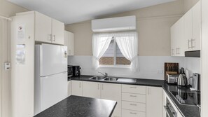 A modern kitchen with white cabinets, a black countertop, a built-in stove, and a window with sheer curtains. - Fishos Hangout 1 - Unit 1 Sinclair St 21 (BERMAGUI)