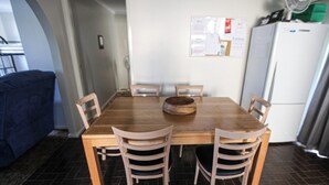 Comedor con mesa de madera y cuatro sillas. Hay un tablón de anuncios en la pared y un refrigerador al fondo.