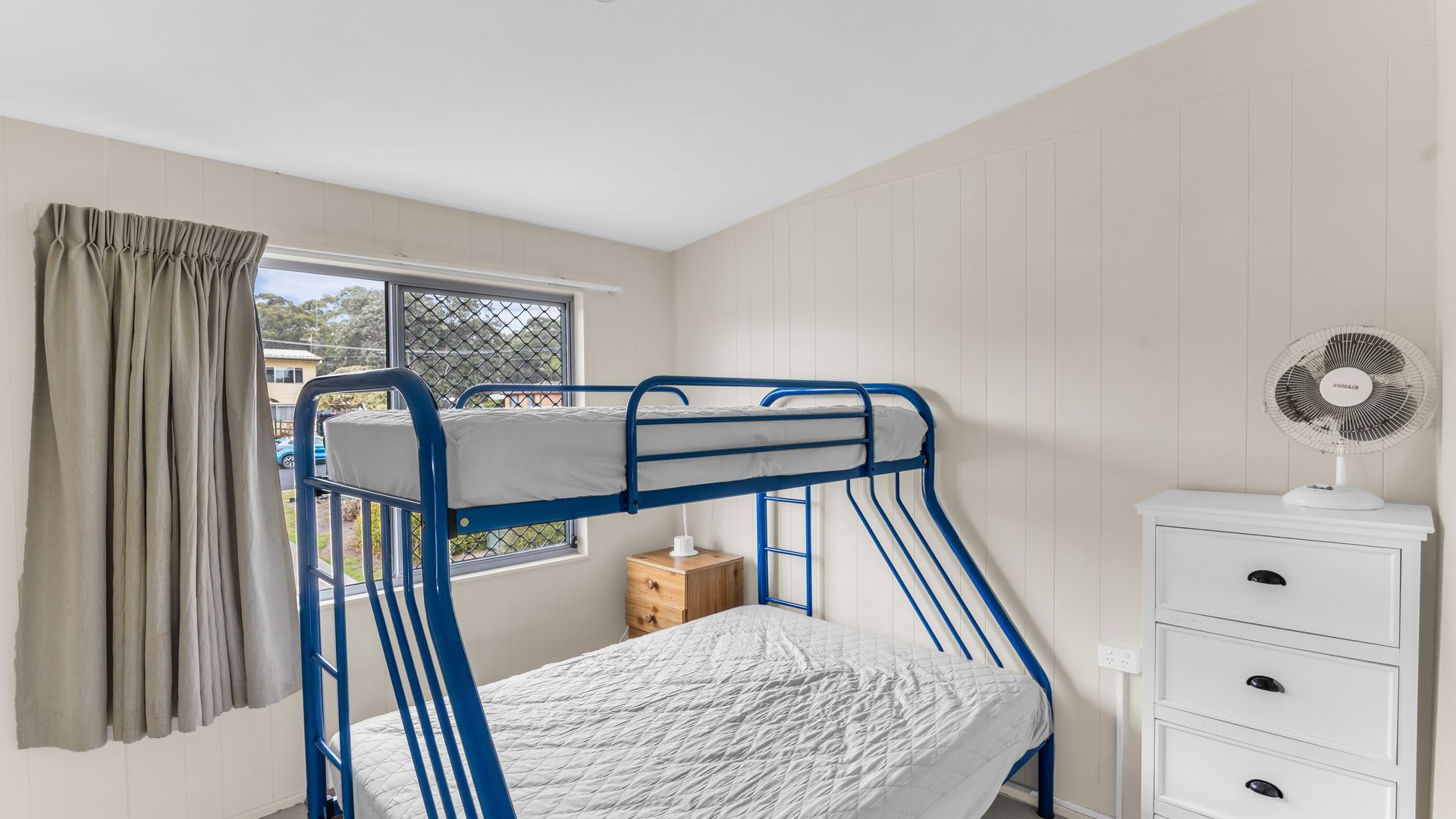 A bunk bed room with a window, a small wooden nightstand, and a white dresser with drawers.