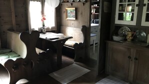 Un interior rústico de madera con una mesa de comedor y bancos, una zona de cocina con armarios y una ventana con cortinas.