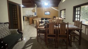 A dining area with a wooden table and chairs, a sofa, and a television in the background. - Recanto do Vale (PIRACAIA)
