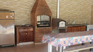 A kitchen with a brick oven, a refrigerator, and a table with a colorful tablecloth. - Recanto do Vale (PIRACAIA)