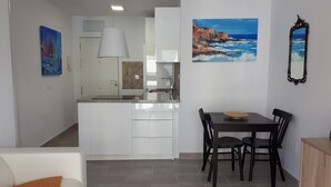 Una cocina moderna con armarios blancos, encimera de granito y una lámpara colgante. Hay una zona de comedor con una mesa y sillas negras, y un cuadro de una escena costera en la pared.