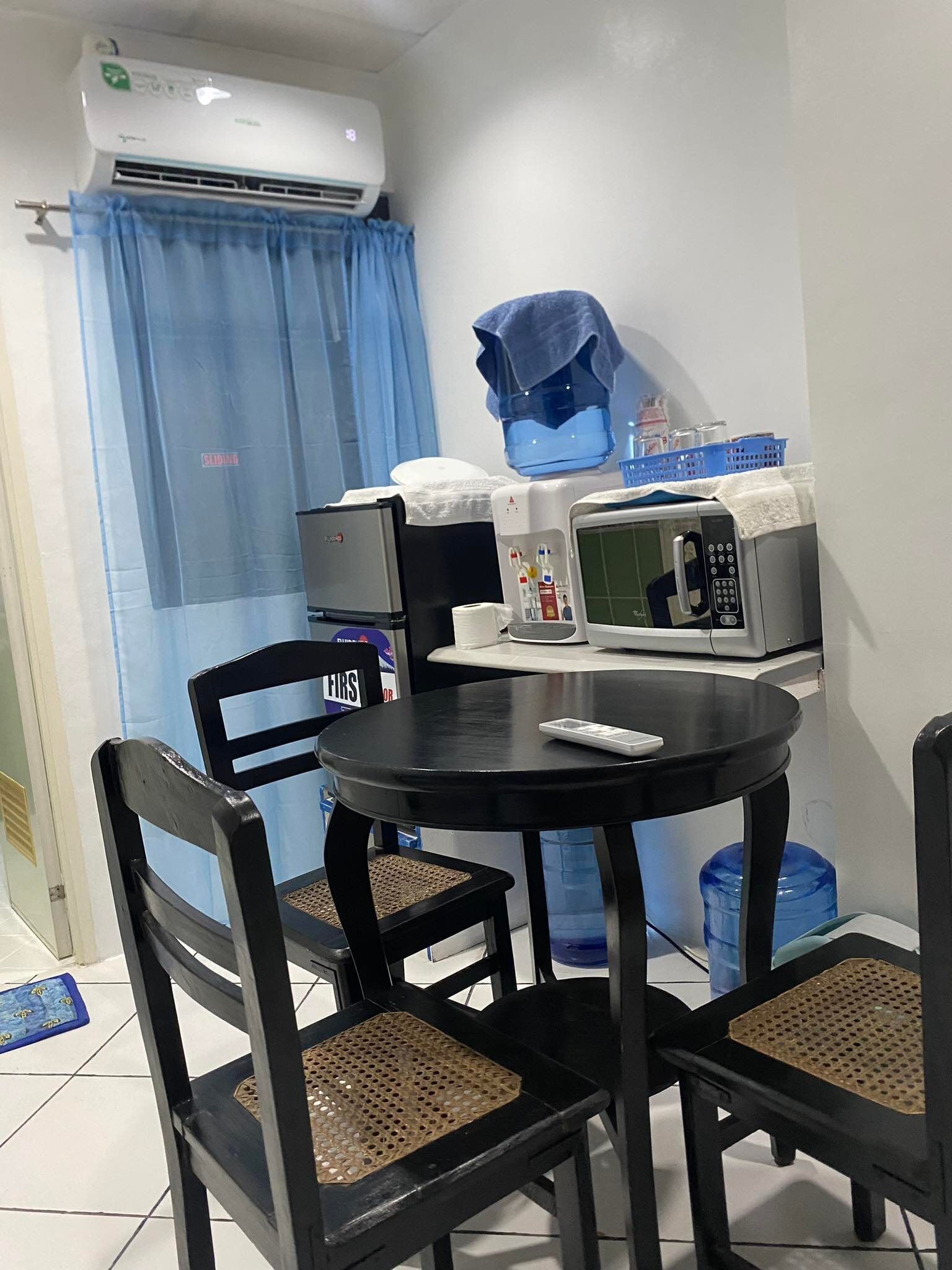 A small kitchenette with a microwave, toaster, and refrigerator. There is a round table with four chairs, and a blue curtain is partially drawn.