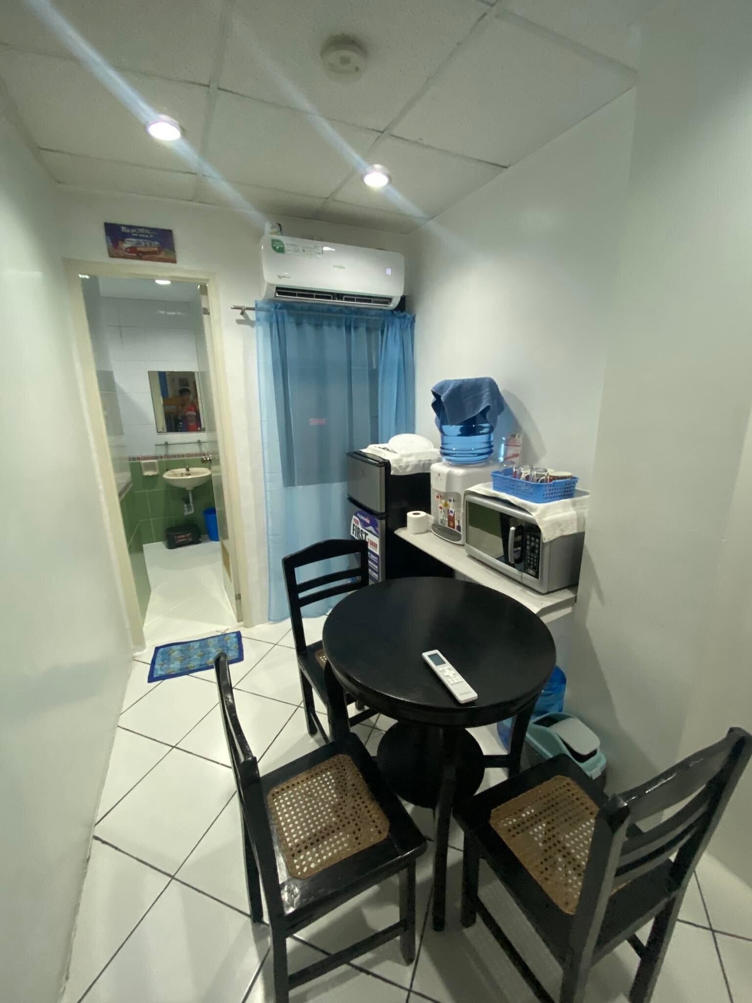 Una habitación pequeña y funcional con una mesa redonda y cuatro sillas, un equipo de aire acondicionado, un microondas y un dispensador de agua.