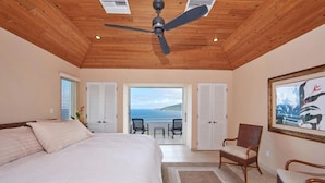 Un dormitorio con cama, ventilador de techo, ventana grande y vista al océano.