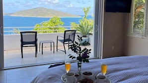 Un dormitorio con una cama grande, un balcón con sillas, televisión y vista al mar.