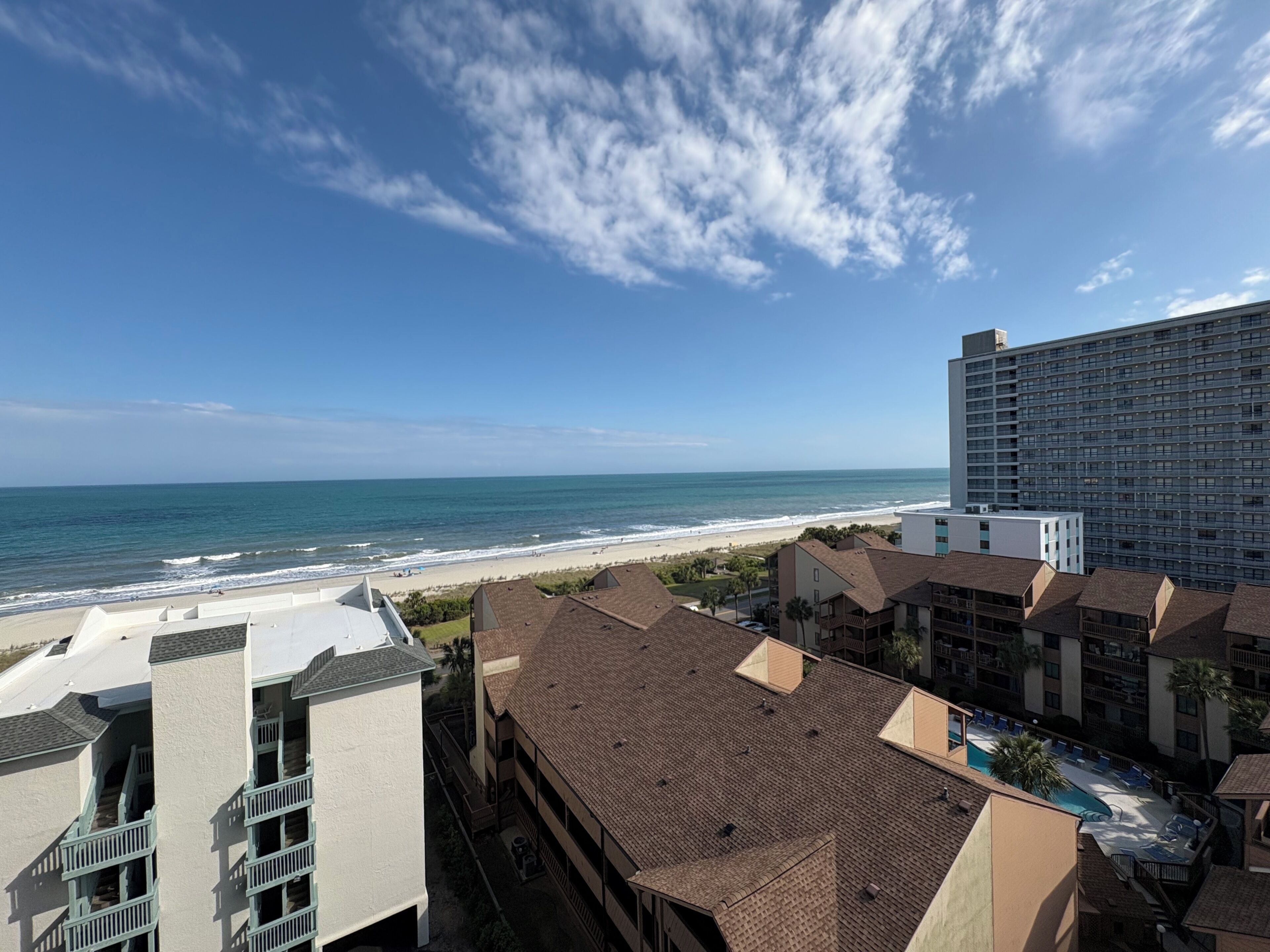 A coastal hotel with a view of the beach and ocean, featuring multiple-story buildings with balconies and a swimming pool.