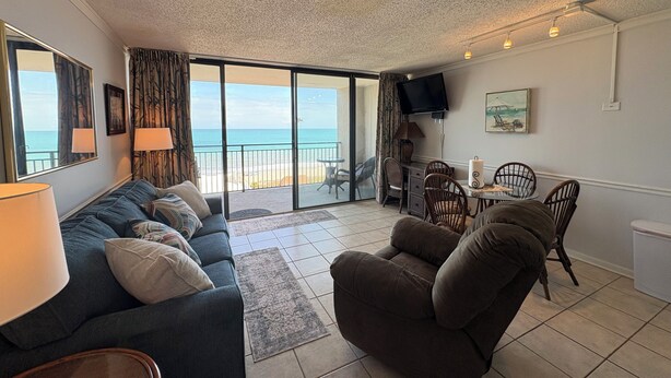 A living room with a sofa, armchair, and a dining table with chairs. There is a TV mounted on the wall, a window with curtains, and a view of the ocean through a sliding door. - Oceanfront Condo in Myrtle Beach  (Myrtle Beach)