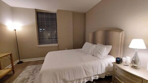 Un dormitorio con cama, mesita de noche, lámpara y ventana con persianas.
