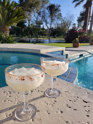 Two margaritas on a poolside table with a pool and greenery in the background.