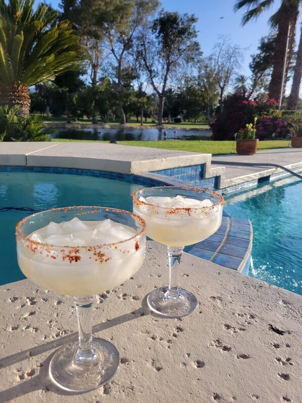Dos margaritas sobre una mesa junto a la piscina, con una piscina y follaje al fondo.