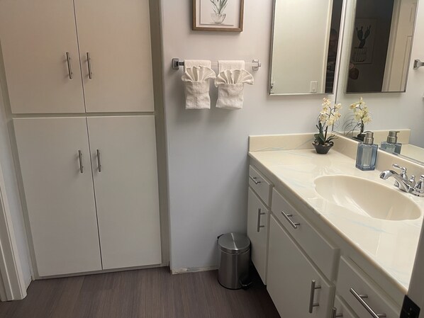 A bathroom with a white sink, mirror, and cabinets. There are towels hanging on a rack and a trash can.