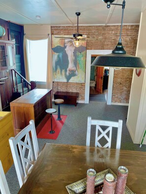 Un coin repas avec une table en bois, des chaises blanches, un tapis rouge et un tableau représentant une vache accroché au mur.
