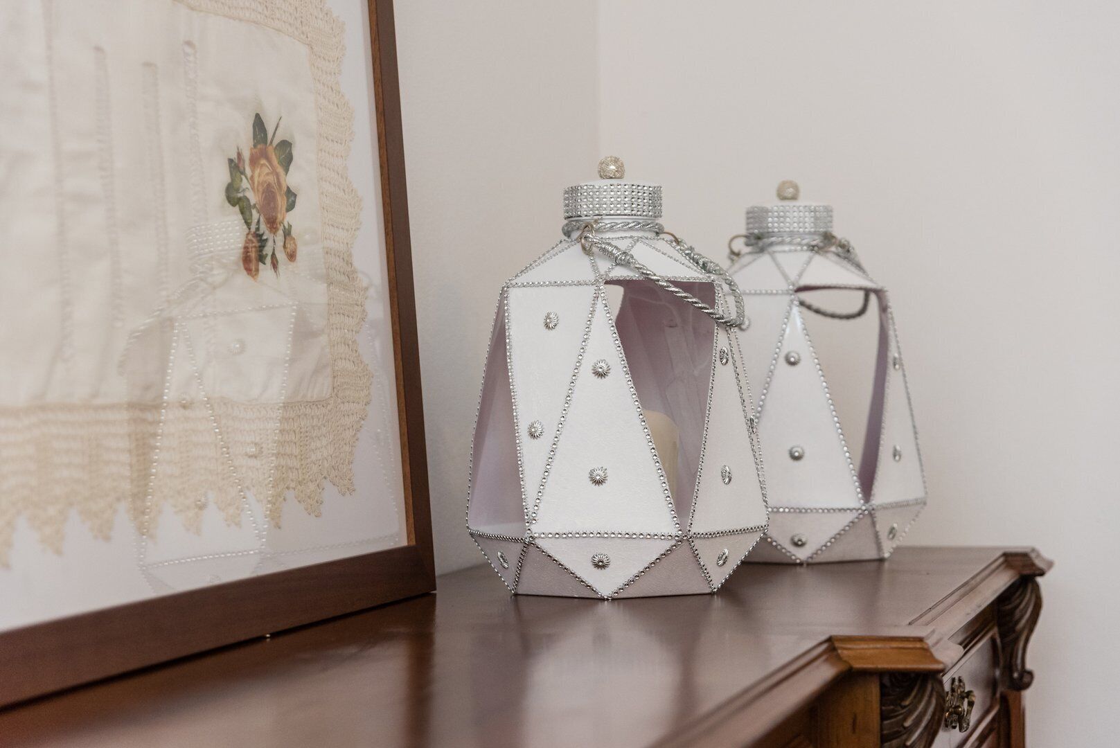 Two silver decorative containers with chain straps and a small knob on top, placed on a wooden surface next to a framed artwork.