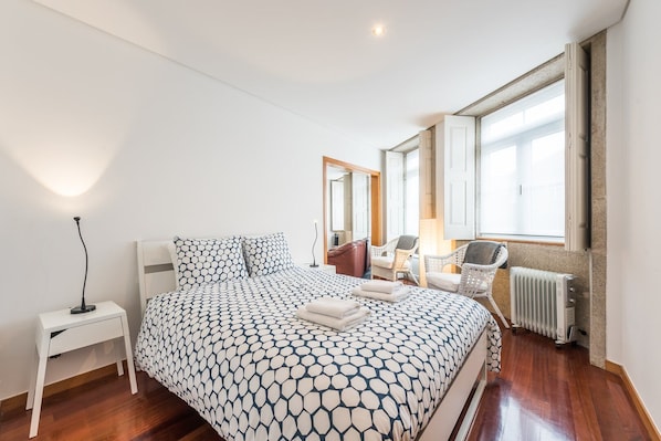 A bedroom with a bed, bedside table, chairs, a radiator, and a window.
