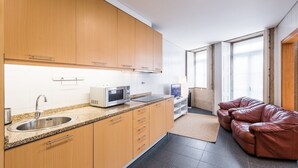 A modern kitchen with wooden cabinets, a stainless steel sink, a microwave, and a flat-screen TV. There is a leather couch and a window with a view.