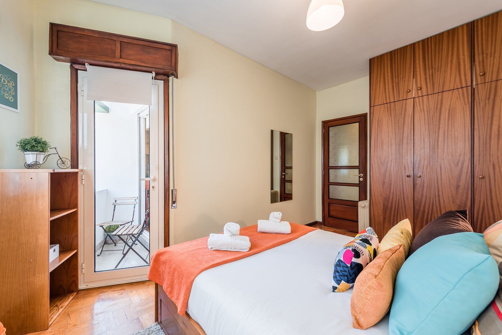 Un dormitorio con cama, armario, puerta que da a un balcón y una mesita con una planta.