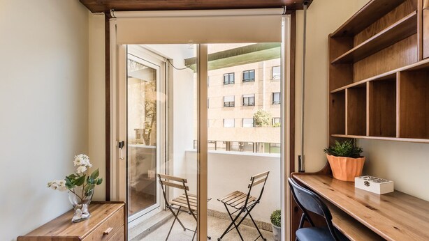 Un petit appartement moderne doté d’une porte-fenêtre coulissante donnant sur un balcon. À l’intérieur, on trouve un bureau en bois, une chaise, une petite bibliothèque et une plante en pot.