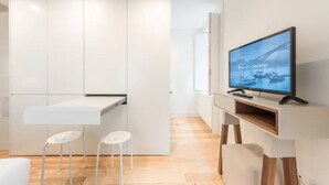 A modern, minimalist room with a flat-screen TV mounted on a wooden stand, a white desk with two stools, and a large wardrobe.